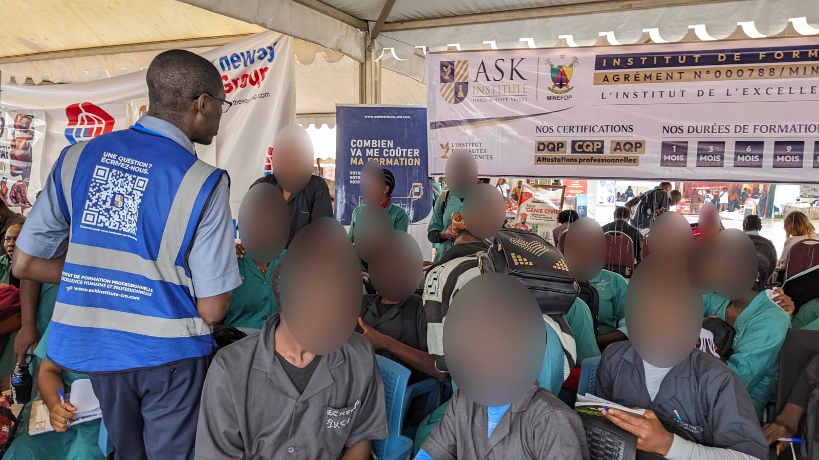 Sur le stand de ASK au CAMBE Ouest 2026, un membre de l’équipe en gilet échange avec un groupe d’élèves, sous la banderole de l’Institut indiquant son agrément MINEFOP, ses certifications et ses durées de formation.