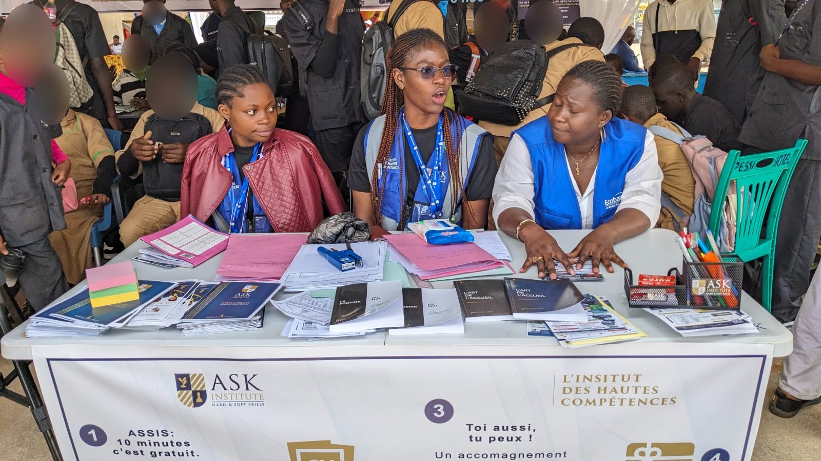 Trois personnes, dont une membre de l’équipe de ASK en gilet de l’Institut, reçoivent les visiteurs à la table du stand couverte de brochures et de formulaires ; des élèves attendent derrière elles.