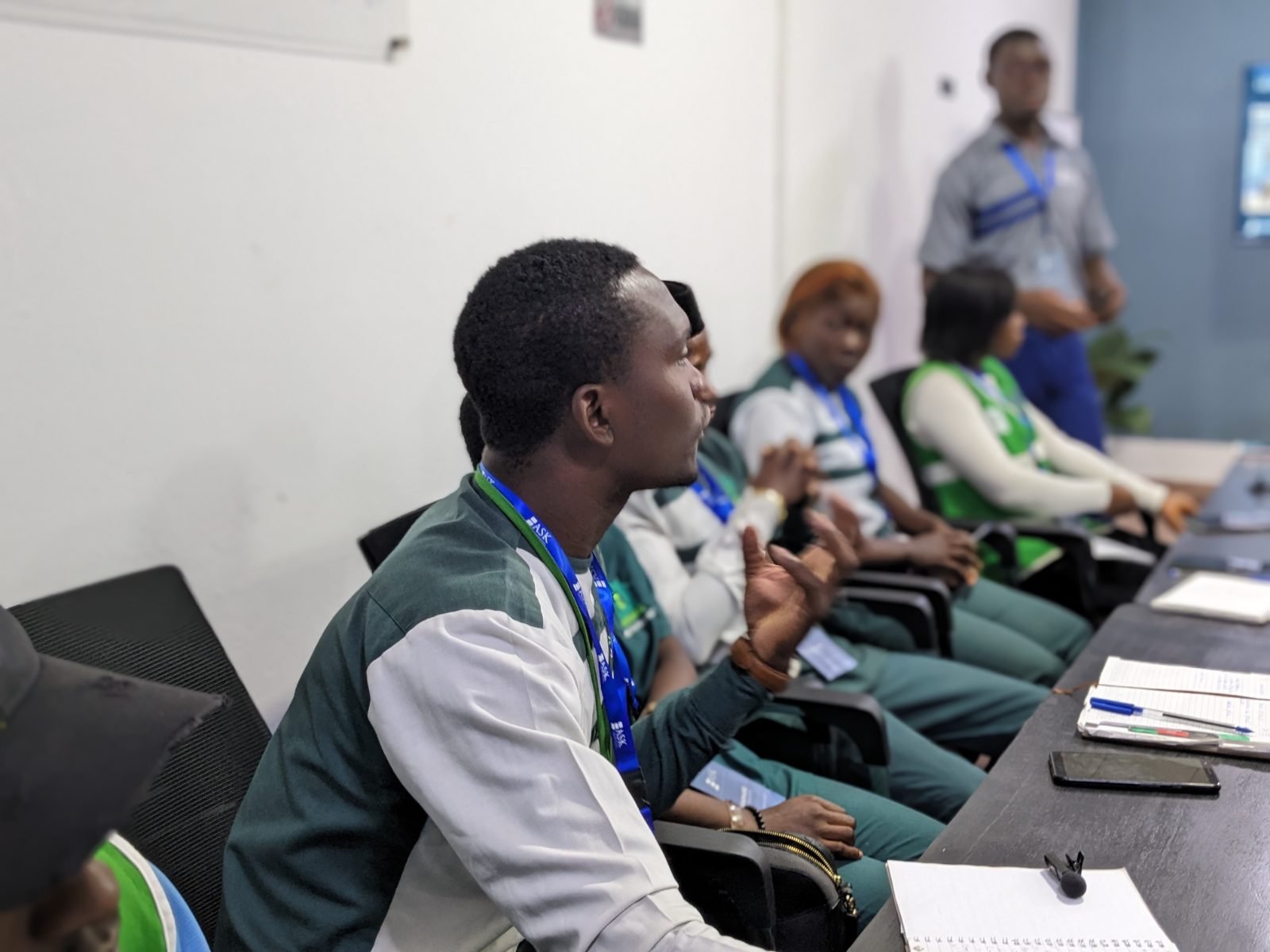 Un participant s’exprime en faisant un geste de la main ; ses collègues l’écoutent ; un membre de l’équipe de ASK Institute se tient debout au fond de la salle.