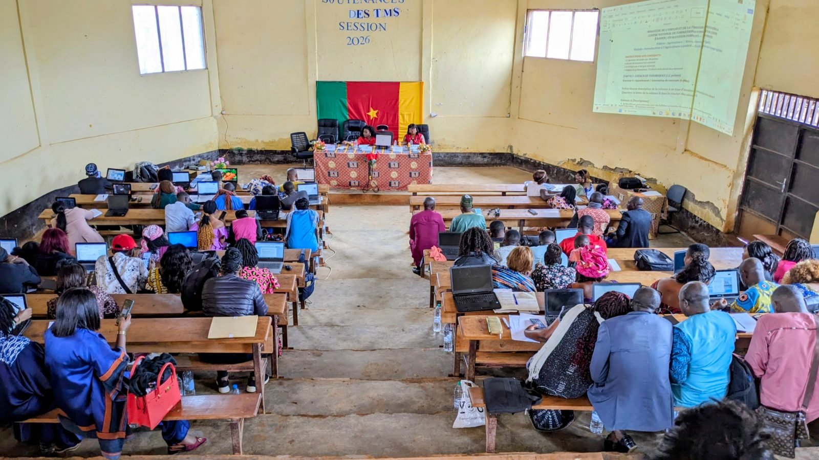 Vue d’ensemble de la salle : les participants, sur leurs ordinateurs portables, font face à la table officielle, devant un drapeau du Cameroun.
