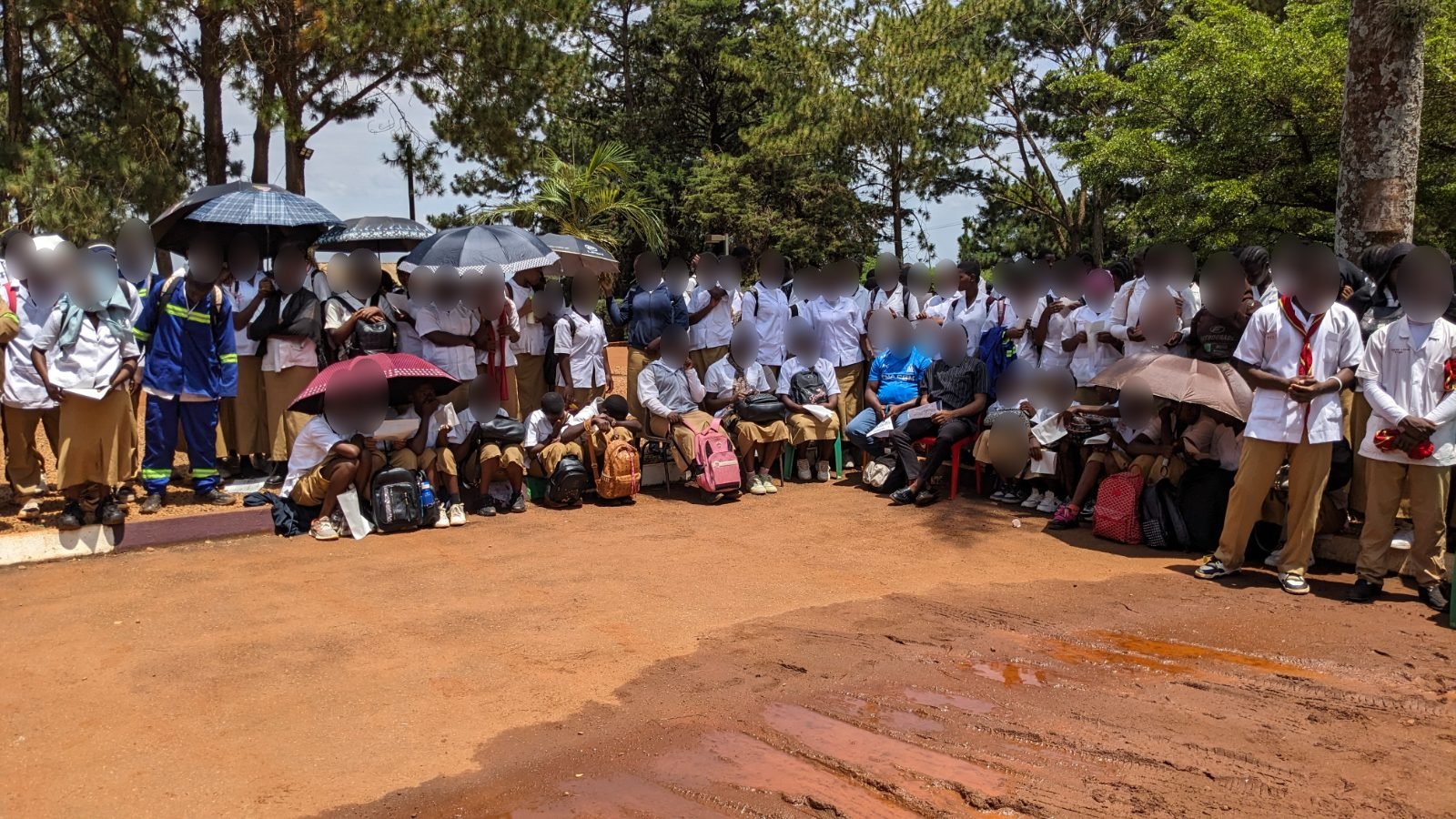 Des élèves en uniforme blanc et beige, debout et assis, certains abrités sous des parapluies, assistent à la cérémonie dans la cour du lycée.