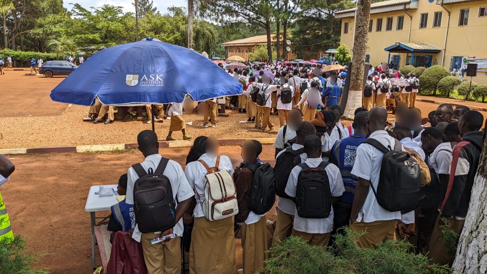 Dans la cour du Lycée Technique de Bafoussam, sous le parasol bleu de ASK Institute, des élèves en uniforme se pressent à la table du stand ; au fond, la cérémonie de la journée se déroule devant le bâtiment administratif.