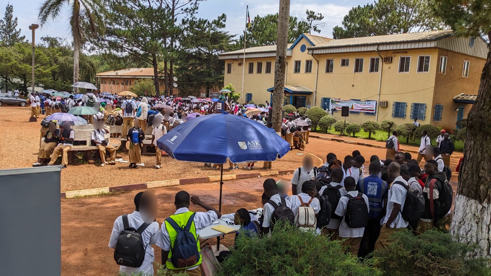 Sous le parasol de ASK Institute, des élèves entourent la table du stand pendant que la cérémonie se poursuit dans la cour du lycée.