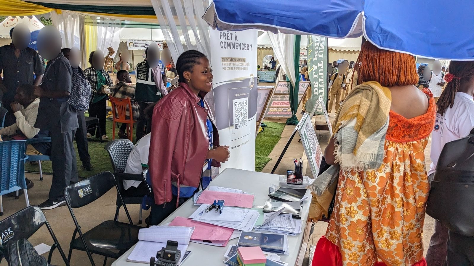 Une membre de l’équipe de ASK accueille une visiteuse à la table du stand, devant le kakémono « Prêt à commencer ? Orientation gratuite, candidature accompagnée ».