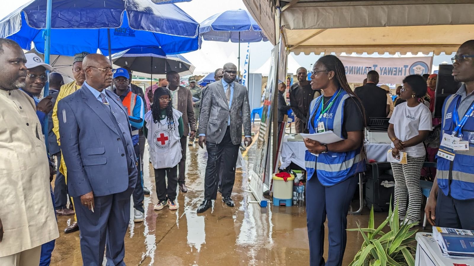 Sous la pluie, S.E. Monsieur le Gouverneur de la Région de l’Ouest et son état-major visitent le stand de ASK ; la Directrice Académique de l’Institut, documents en main, leur présente les activités de l’Institut.