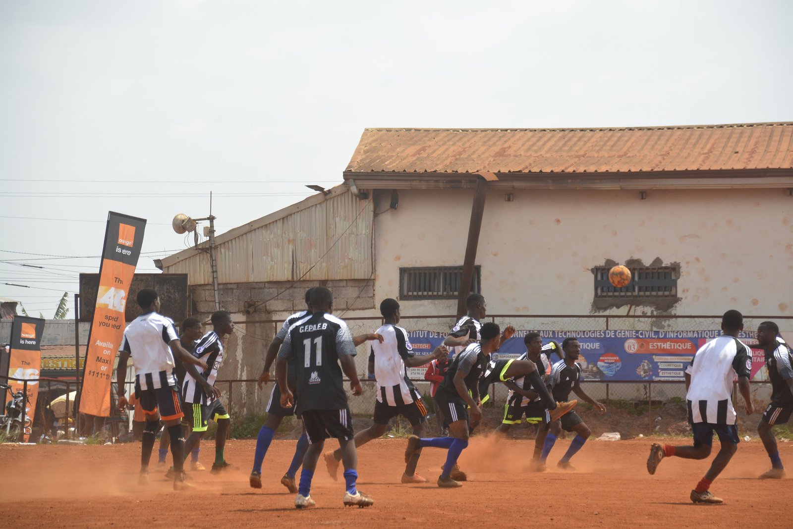 Phase de jeu d’un match de football, devant un bâtiment au bord du terrain.