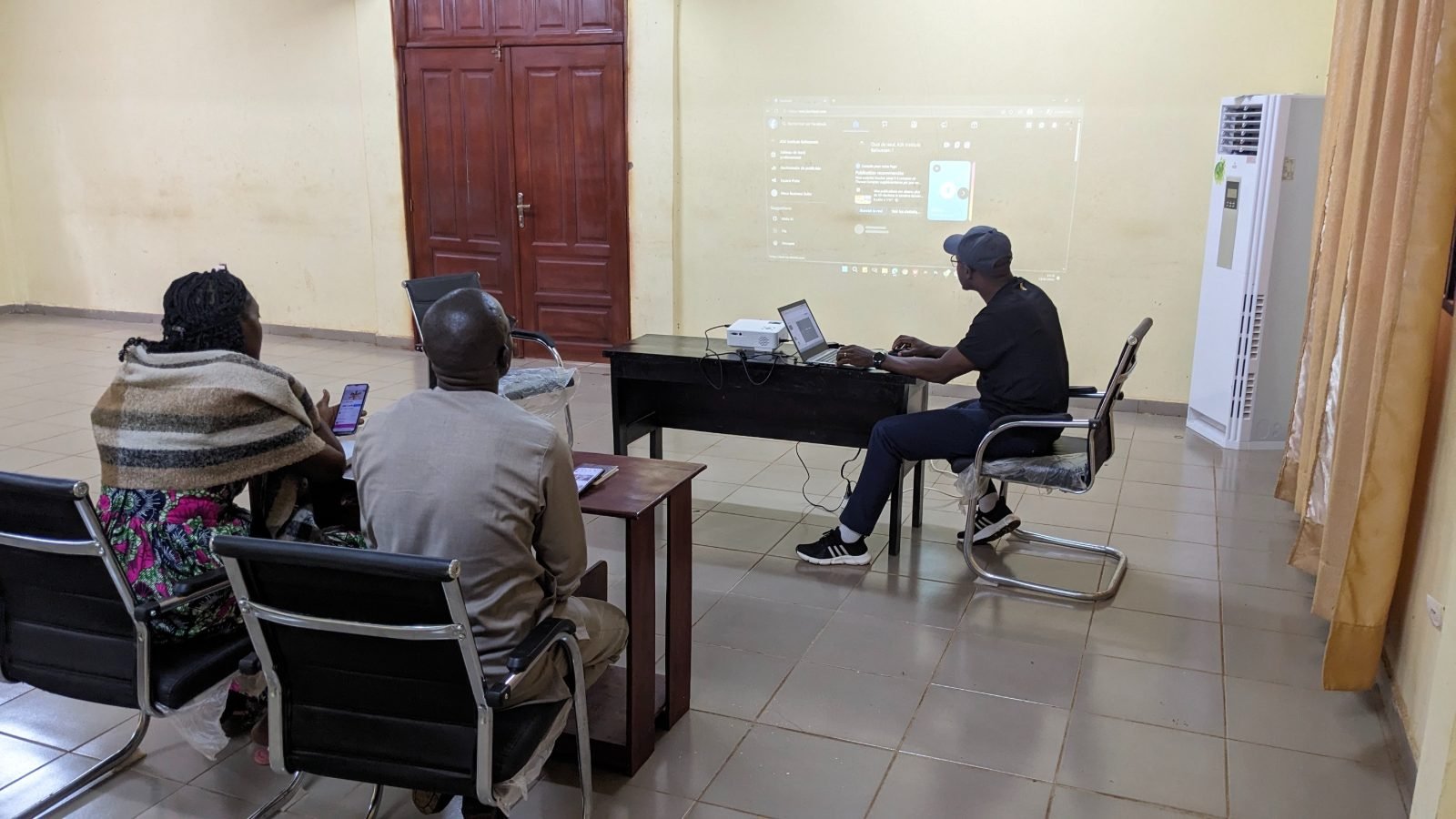 Formation des administrateurs du CIOP Bafoussam : un formateur, assis devant un ordinateur portable et un vidéoprojecteur, projette au mur l’interface de gestion d’une page professionnelle sur les réseaux sociaux ; deux participants suivent la démonstration, l’une téléphone en main.
